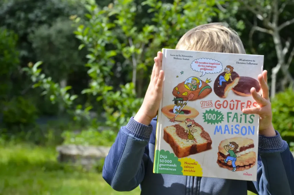 Petit garçon tenant devant sa tête le livre "Mes goûters 100% faits maison"